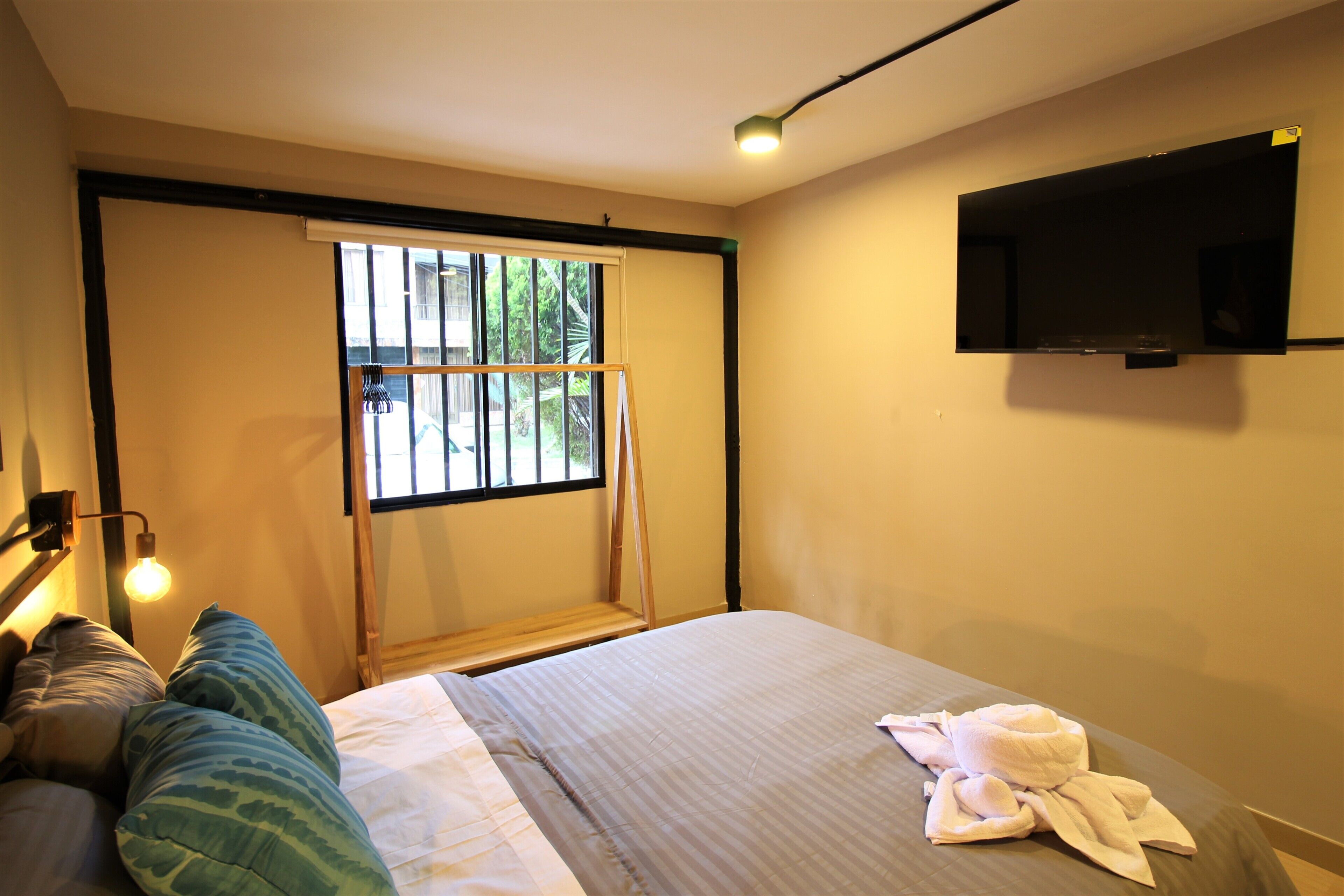 A hotel room with a bed, a TV mounted on the wall, a window with a view of greenery, and a towel on the bed.