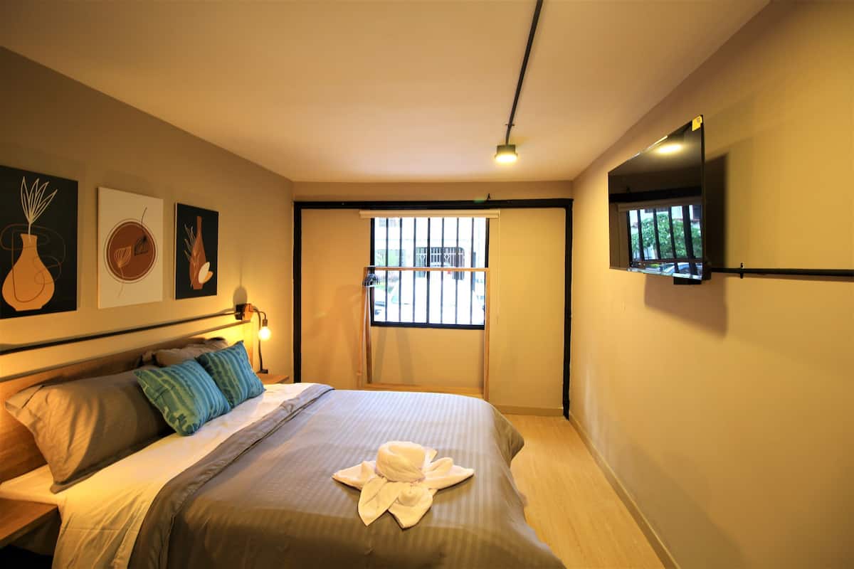 Une chambre d’hôtel avec un lit, une table de chevet, un téléviseur fixé au mur et une fenêtre avec des stores.