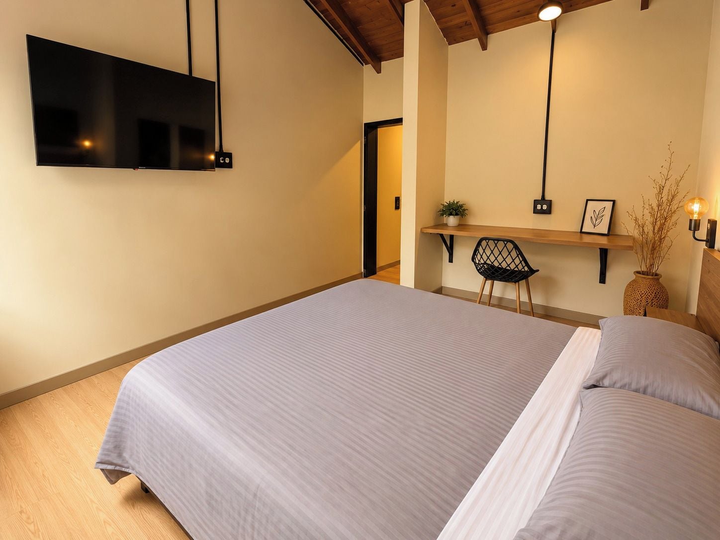 A bedroom with a bed, a TV mounted on the wall, a desk with a chair, and a bedside table with a lamp.