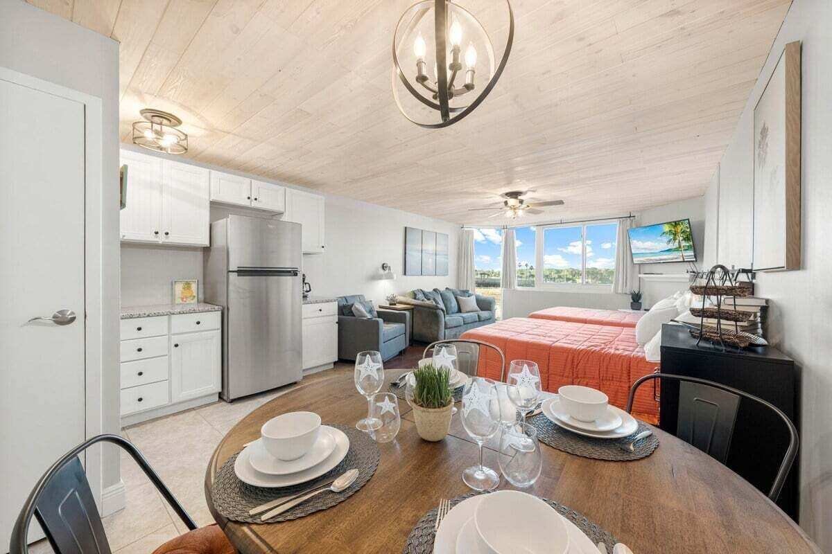 A modern kitchen with white cabinets and a stainless steel refrigerator. There is a dining area with a wooden table set for four, and a bed with an orange bedspread in the background.