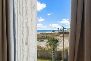 A beach view through a window with curtains, including a sandy beach, palm trees, and a clear sky.