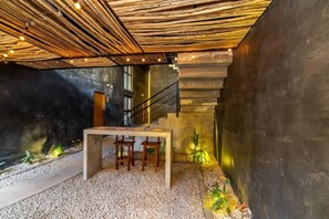 Stairs lead to an upper level, with a table and chairs below, surrounded by potted plants and a textured wall.