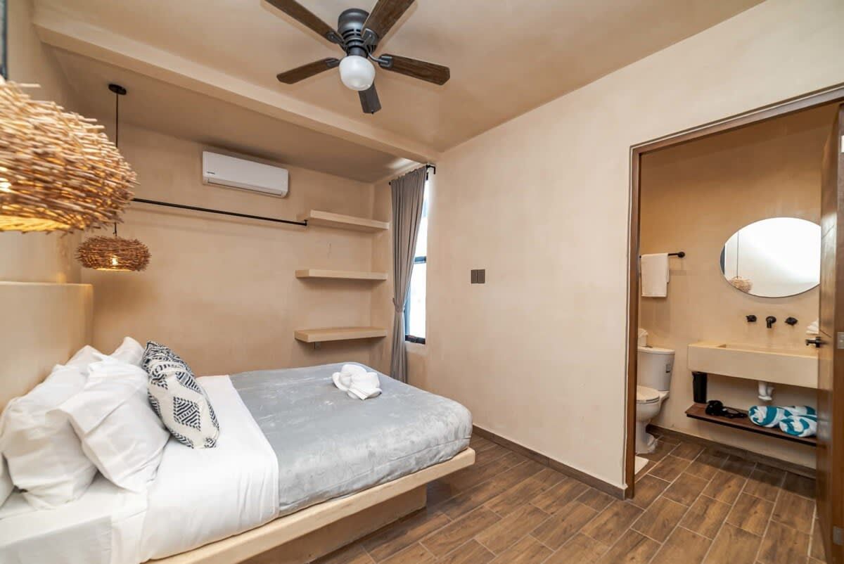 A bedroom with a bed, a ceiling fan, a hanging light fixture, a built-in shelf, a window with curtains, and a bathroom with a toilet, sink, and mirror.