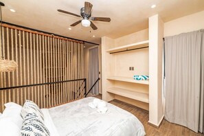 Une chambre d’hôtel moderne équipée d’un lit, d’un ventilateur de plafond, d’une cloison en lattes de bois, d’une étagère intégrée et d’un rideau.