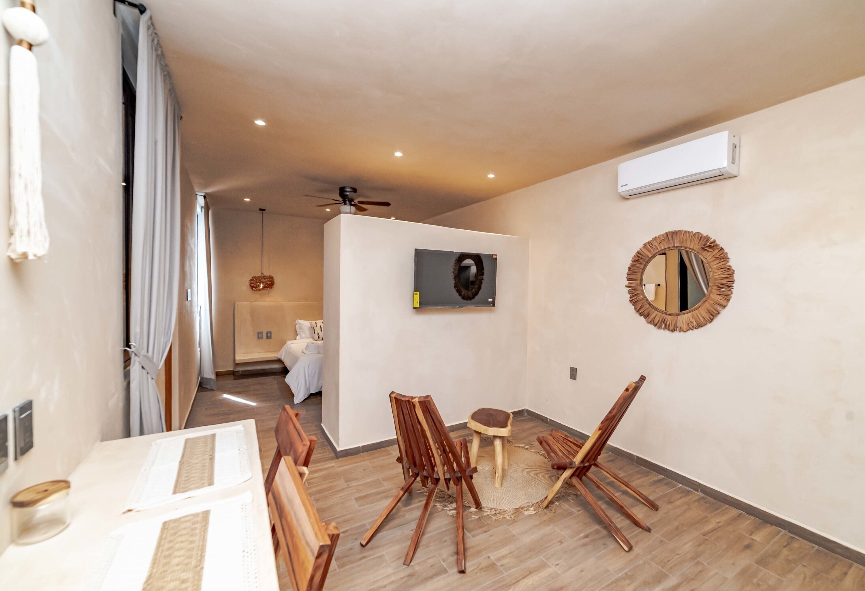A room with a wooden floor, a ceiling fan, an air conditioning unit, a television, a mirror, and a dining area with chairs and a table.