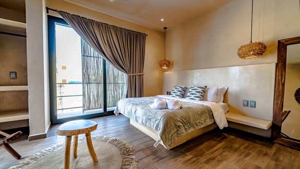 Una camera da letto con un letto, uno sgabello rotondo in legno, un tappeto tessuto, una ventola a soffitto e un'ampia finestra con tende.