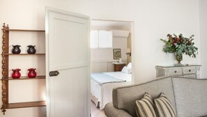A hotel room with a bed, a sofa, a wooden shelf with decorative items, and a bedside table with a vase of flowers.