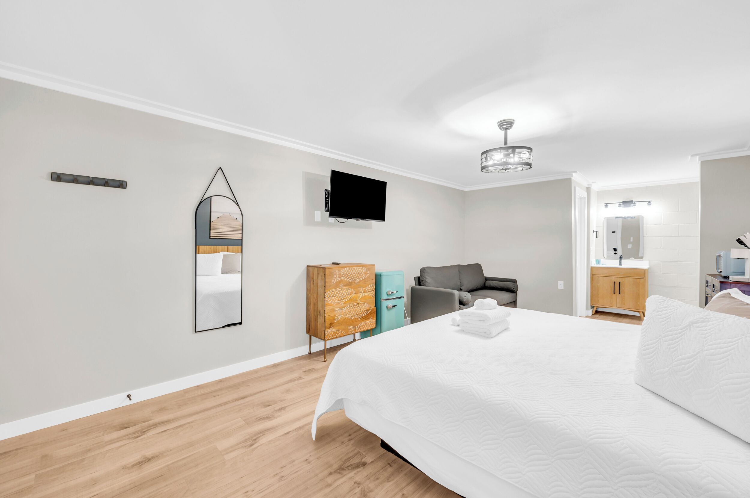 A modern hotel room with a large bed, a flat-screen TV mounted on the wall, a wooden cabinet, a gray armchair, and a small desk with a lamp.