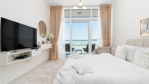 A modern hotel room with a large bed, a flat-screen TV, a seating area with chairs, and a balcony with a view. - Elegant Studio at Damac Hills Carson B (Dubai)