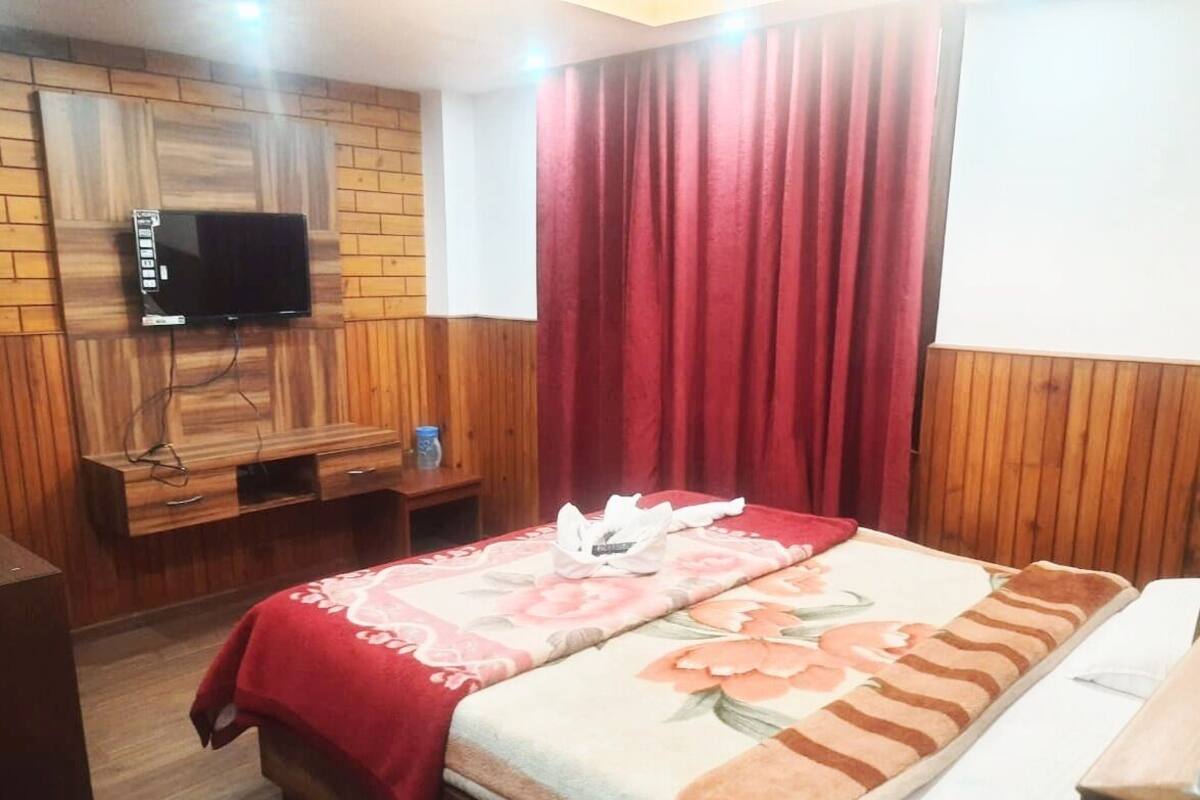 A hotel room with a wooden paneled wall, a flat-screen TV mounted above a wooden cabinet, a bed with floral bedding, and a red curtain.