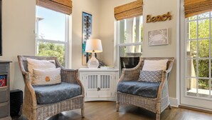 A room with two wicker chairs, a lamp, a wall sign, and a view of greenery outside.