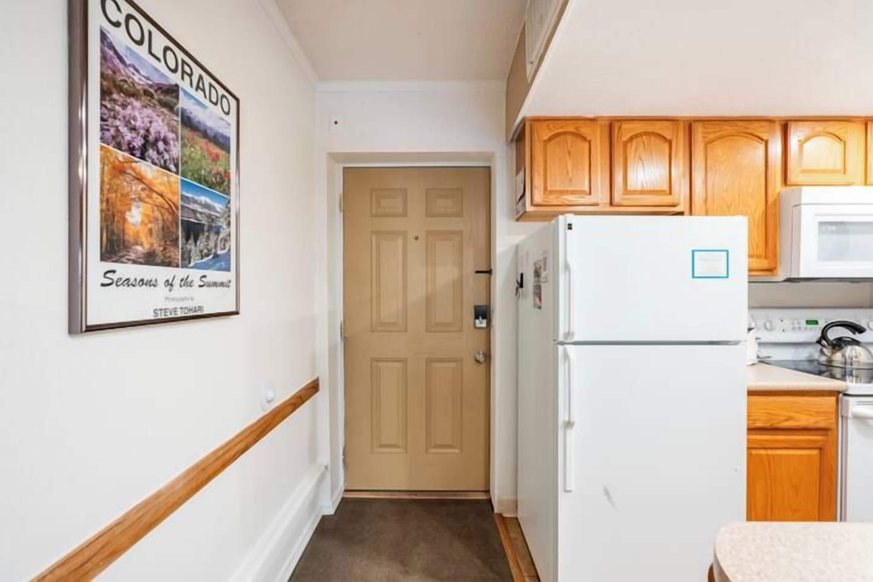 A narrow hallway leading to a door, a white refrigerator, wooden cabinets, and a microwave.
