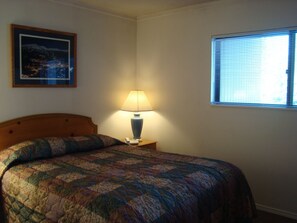 A neatly made bed with a patterned quilt, a bedside lamp, a framed picture on the wall, and a window with blinds.