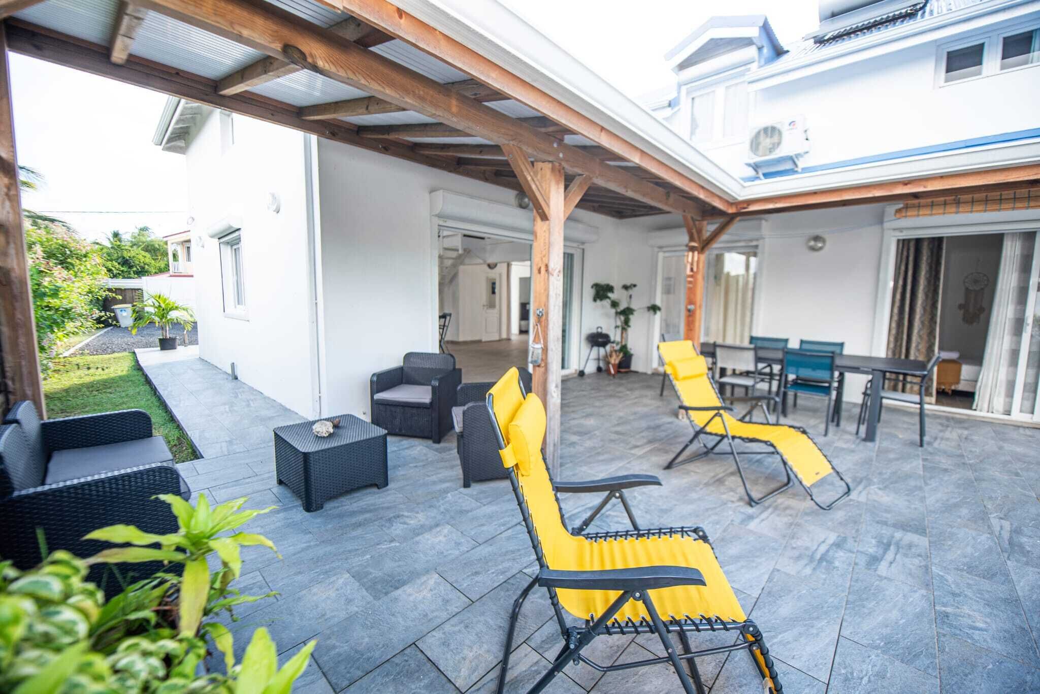 Un'area patio esterna con posti a sedere, inclusi divani neri e poltrone gialle, e una zona coperta con una pergola.