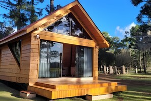 A wooden cabin with a large glass door and windows, set in a wooded area. - Cabana Pequeno Bosque Com Vista - o Rio, à 15 km da cidade (São Joaquim)