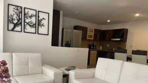 A modern living room with a white sofa, a coffee table, and a potted orchid. There is a kitchen in the background with cabinets and appliances. - Exclusive condominium ideal for families with babies and pets. (Envigado)