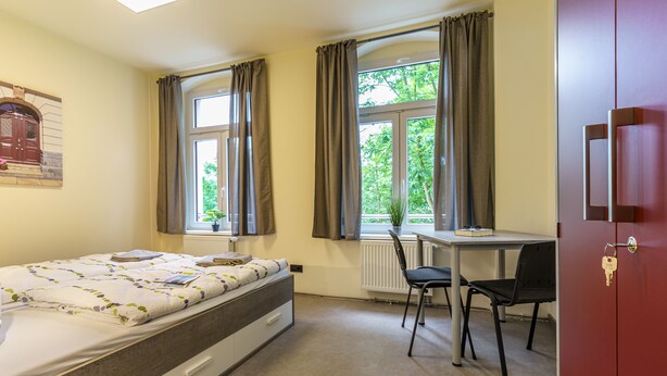 A bedroom with a bed, a desk, a chair, and a window with curtains. - Hostel Gleisbett (Annaberg-Buchholz)