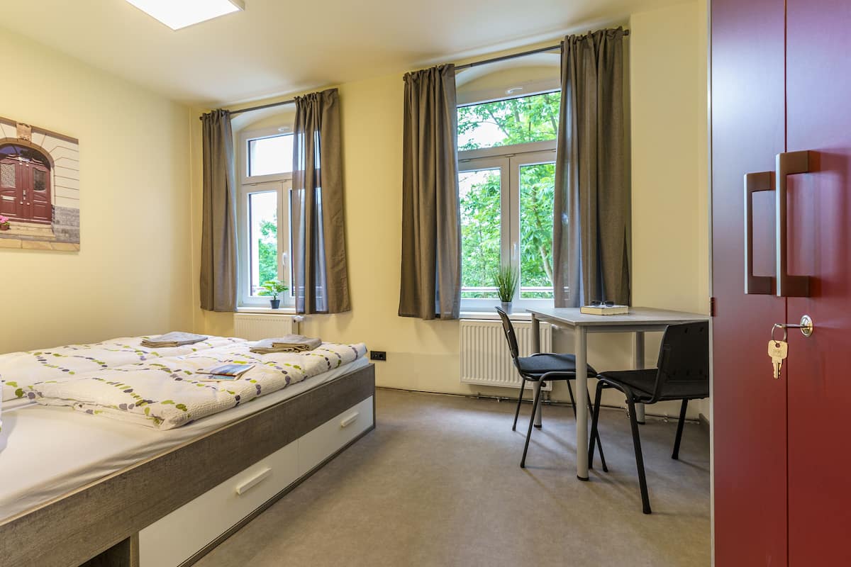 A bedroom with a bed, a desk, a chair, and a window with curtains.