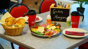 A breakfast table with a variety of food items, including bread, cheese, meats, and fruits. There are red and green cups, a basket of bread, and a sign that reads "Guten Morgen". - Hostel Gleisbett (Annaberg-Buchholz)