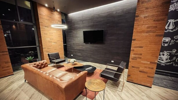 A modern living room with a brown leather sofa, a coffee table, and a small round stool. There is a flat-screen TV mounted on the wall and a large window providing natural light.