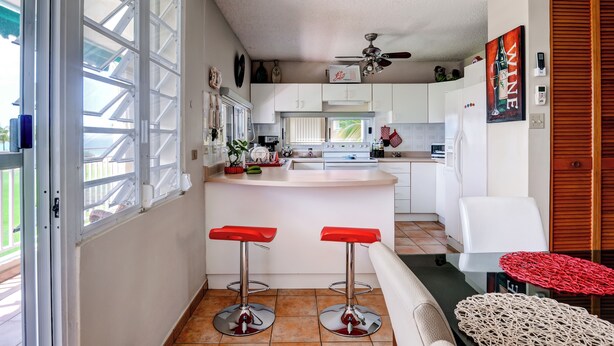 Una cocina compacta con armarios blancos, una isla central, dos taburetes rojos y un ventilador de techo.