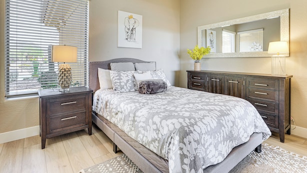 Une chambre à coucher comprenant un lit, des tables de chevet, une commode, un miroir et une fenêtre avec des stores.