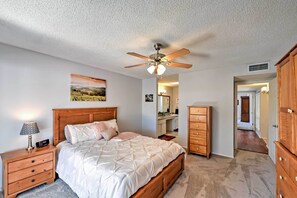 Une chambre à coucher comprenant un lit, une table de chevet en bois, une commode, un ventilateur de plafond et un couloir menant à une autre pièce.