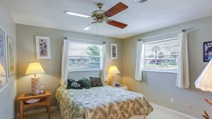 Un dormitorio con cama, dos mesitas de noche, dos lámparas, un ventilador de techo y ventanas con cortinas.
