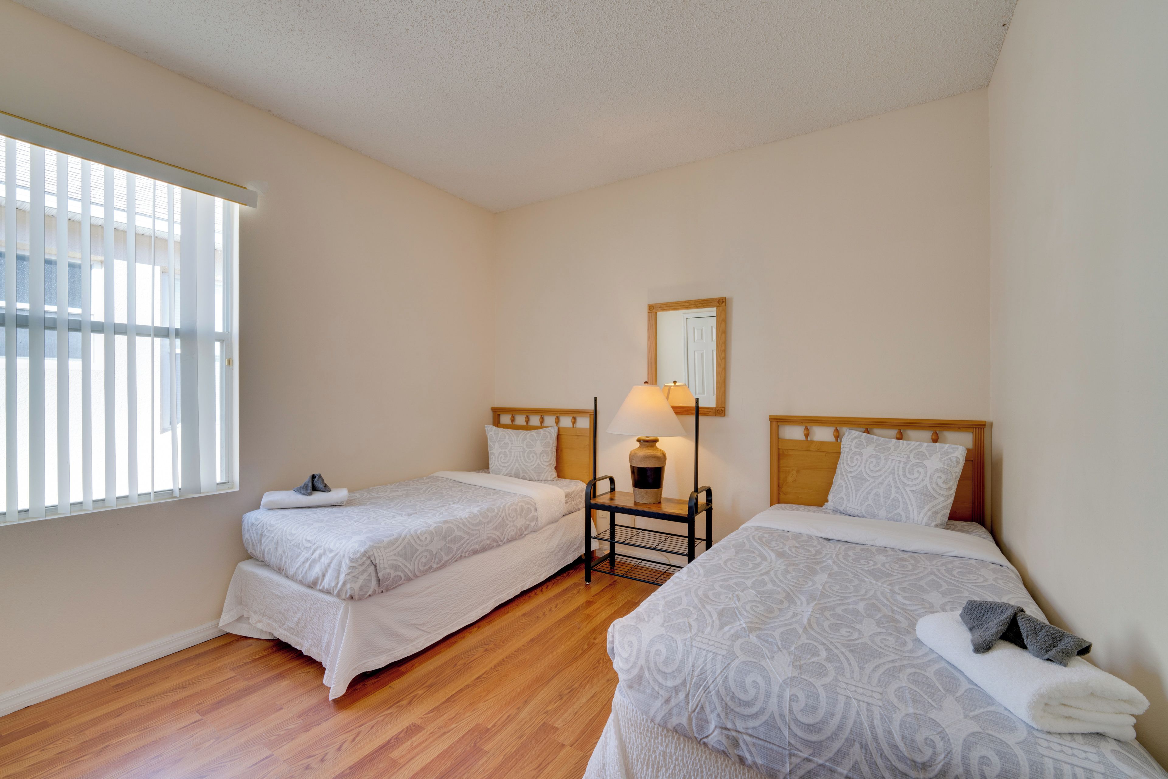 A bedroom with two single beds, a nightstand with a lamp, a mirror, and a window with blinds.