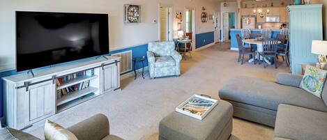 A living room with a large flat-screen TV, a sofa, ottoman, and a dining area with chairs and a table.
