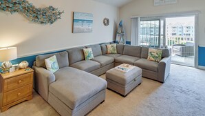A living room with a sectional sofa, a wooden nightstand, and a large window with a view of the beach and buildings. - Pool Access & Balcony: Modern Myrtle Beach Condo (Myrtle Beach)