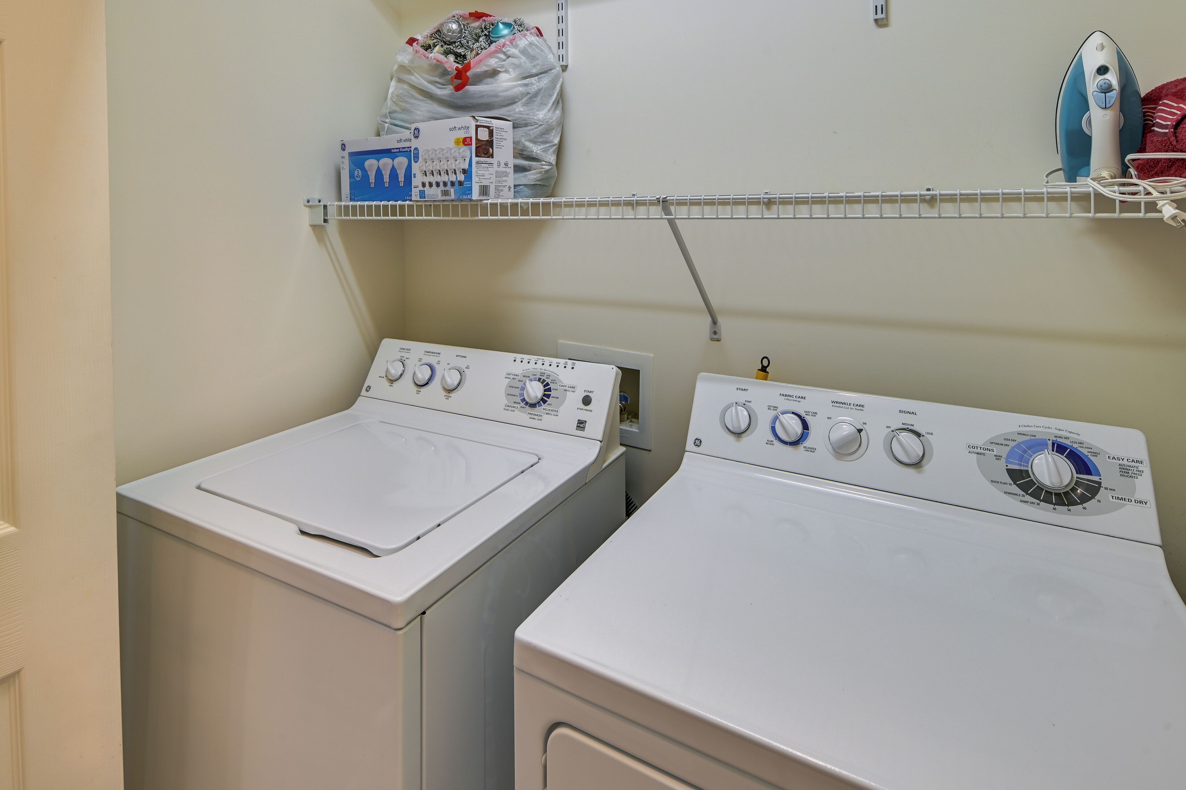 Two front-loading washing machines in a laundry area with a shelf above containing a bag of laundry and a box of detergent.