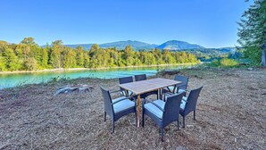 An outdoor seating area with a table and chairs, surrounded by trees and a view of a lake and mountains. - Dreamy Riverside Cabin With Fireplace & Grill (Concrete)