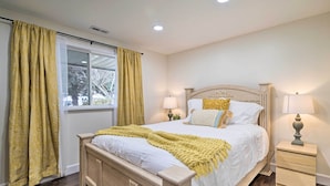 Une chambre à coucher avec un lit en bois, deux tables de chevet avec des lampes, une fenêtre avec des rideaux jaunes et une vue sur les arbres à l’extérieur.