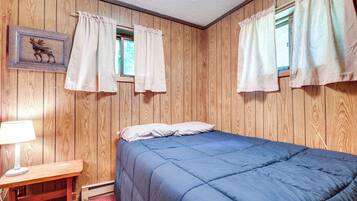 Une chambre avec un mur lambrissé en bois, un lit avec une literie bleue, une table de chevet avec une lampe, et deux fenêtres avec des rideaux.