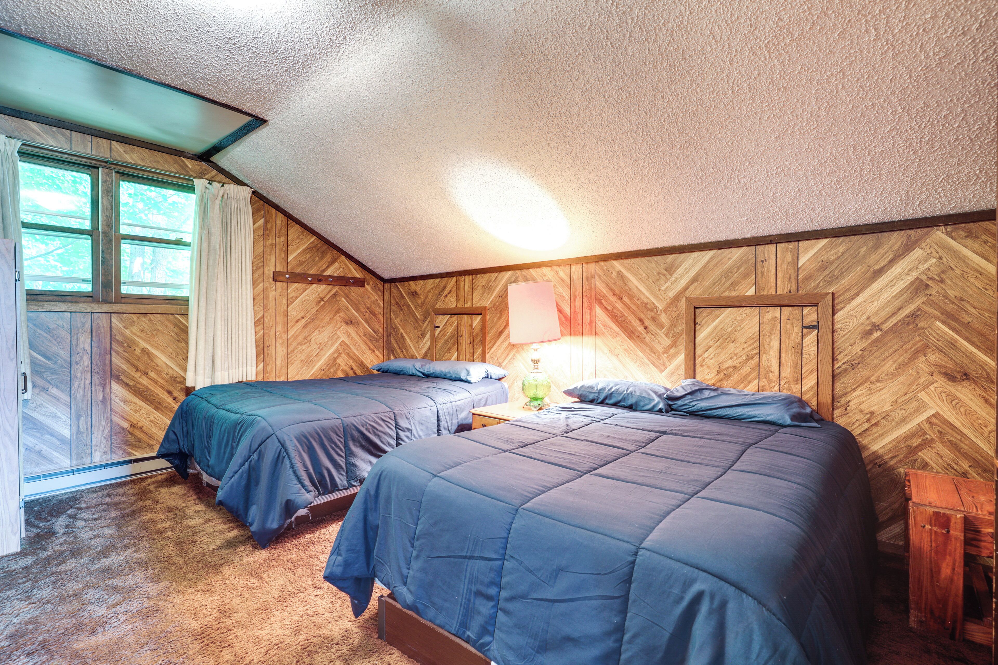 A bedroom with two beds, wooden walls, a window with curtains, and a nightstand with a lamp.