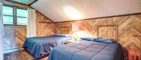 A bedroom with two beds, wooden walls, a window with curtains, and a nightstand with a lamp.