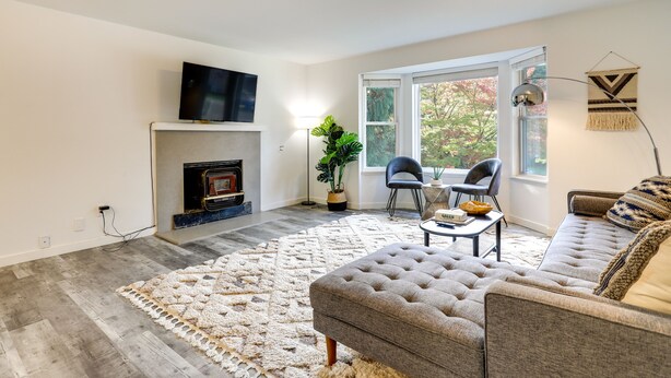 A living room with a fireplace, a television mounted above it, a grey sofa, a coffee table, and a large window providing a view of trees. - Beautiful Kenmore Retreat Near Marina & Beach! (Kenmore)