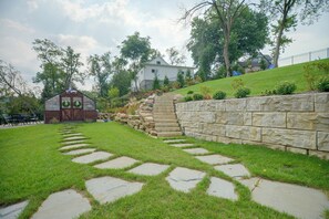 A stone pathway leading to a wooden structure with a roof, surrounded by a well-maintained lawn and a stone retaining wall.