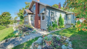 A two-story house with a brick chimney, a front porch, and a well-maintained garden with various plants and flowers. - Vintage Charlevoix Home w/ Deck Near Round Lake! (Charlevoix)