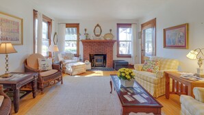 A cozy living room with a fireplace, wicker furniture, and a checkered sofa. - Vintage Charlevoix Home w/ Deck Near Round Lake! (Charlevoix)