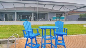 A covered outdoor seating area with blue chairs and a table, a wooden deck, and a building with large windows and a covered roof in the background. - Canal-view Oasis w/ Grill in Cape Coral! (Cape Coral)