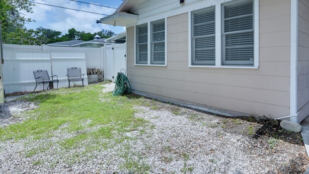 A single-story house with a beige exterior, white trim, and two windows with white shutters. There is a small grassy area with a few chairs and a hose. - Clearwater Vacation Rental w/ Furnished Patio! (Clearwater)