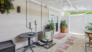 An air conditioning unit, a table, and chairs in a covered outdoor area with a white ceiling and walls, surrounded by potted plants and a gravel pathway. - Clearwater Vacation Rental w/ Furnished Patio! (Clearwater)