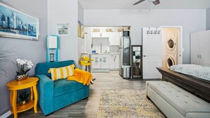 A compact living space with a sofa, a small side table, a bed, a ceiling fan, a kitchen area with cabinets and appliances, and a washing machine. - Clearwater Vacation Rental w/ Furnished Patio! (Clearwater)