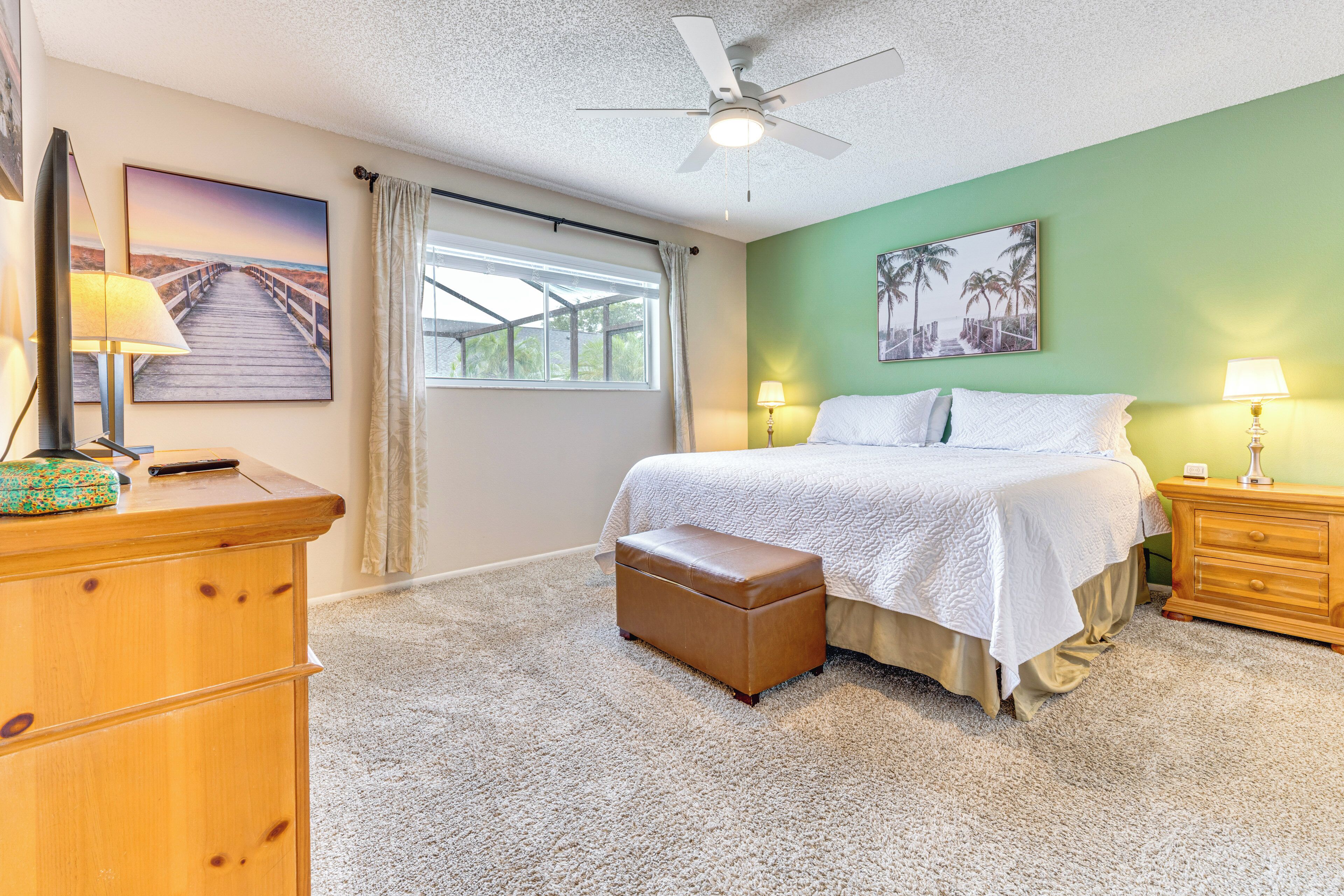 A bedroom with a bed, a ceiling fan, a window with curtains, a dresser, and a nightstand with a lamp.