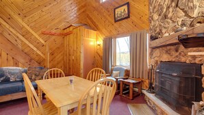 A cozy cabin interior with a wooden dining table, chairs, a sofa, and a stone fireplace.