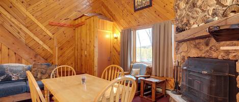 A cozy cabin interior with a wooden dining table, chairs, a sofa, and a stone fireplace.