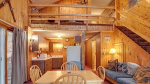 A wooden cabin interior with a dining table, chairs, a sofa, a kitchen area, and a staircase.
