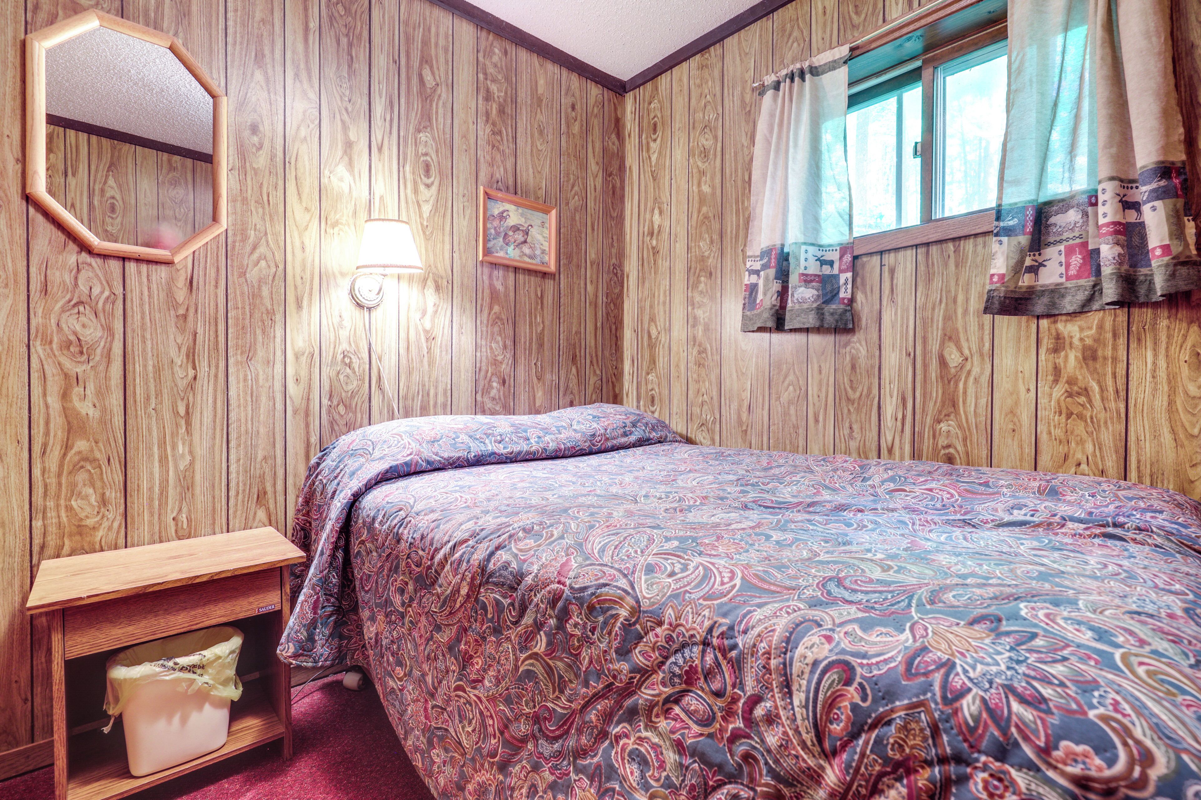 A bedroom with wooden paneling, a bed with a patterned bedspread, a bedside table with a lamp, and a window with curtains.